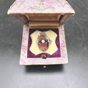May include: A silver-toned ring featuring an elongated, faceted, translucent purple stone with a small, round, white center accent. The ring is displayed inside a hinged, fabric-covered box with a striped exterior and a purple velvet interior.