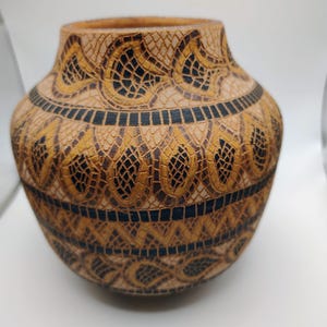 May include: A decorative, handcrafted vase with a textured surface. The vase features a repeating pattern of gold and black geometric shapes. The vase is a light brown color and has a wide, rounded body.
