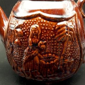 May include: A brown ceramic teapot with a detailed embossed design of people and plants. The teapot has a lid and a spout.