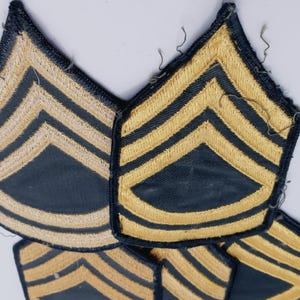 May include: Several vintage military patches with a dark blue background and gold chevrons. The patches are arranged in a cluster, showcasing their design and detail. The patches are likely for a Sergeant rank.