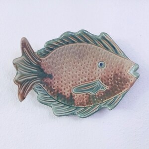 May include: A ceramic fish wall plaque featuring a textured, stippled body in shades of brown and tan. The fins and tail are glazed in a muted sage green with darker brown accents. A single small, dark blue eye is visible on the fish's head.