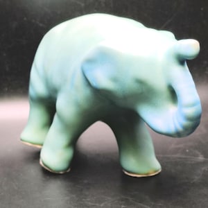 May include: A ceramic elephant figurine featuring a smooth, matte finish with a gradient color scheme transitioning from soft teal to light blue. The elephant is depicted in a walking stance with its trunk curled upward and ears slightly flared.