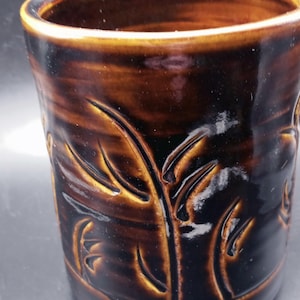May include: A cylindrical ceramic mug with a glossy dark brown glaze. The exterior surface features a carved, repeating leaf or bamboo-like pattern that reveals a lighter golden-brown tone within the recessed grooves of the design.