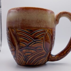 May include: A brown ceramic mug with a textured, leaf-like design. The mug has a light brown rim and a handle.
