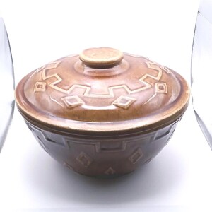 May include: A round, brown ceramic bowl with a matching lid. The exterior of both the bowl and the lid features a raised, geometric pattern consisting of repeating square and diamond shapes embossed into the glazed, glossy brown surface.