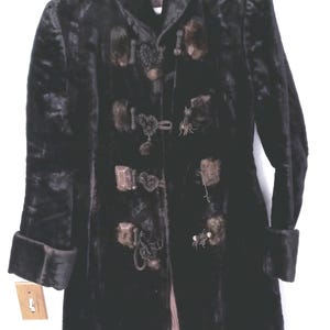 May include: A long, black coat with a high collar and long sleeves featuring folded cuffs. The front closure consists of several decorative toggle-style fasteners with dark brown fur accents and braided cord details. The interior lining is brown.