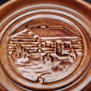 May include: A circular brown ceramic coaster featuring a raised, embossed design of a log cabin nestled among trees. The surface has a glossy, monochromatic finish with concentric circular ridges framing the central cabin illustration.