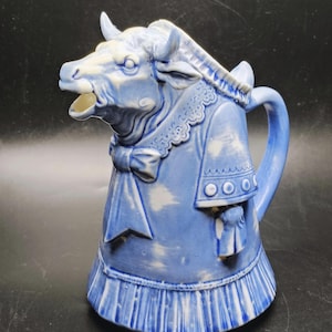 May include: A blue and white ceramic pitcher in the shape of a cow. The cow is wearing a dress with a bow and a ruffled collar. The pitcher has a handle and a spout.