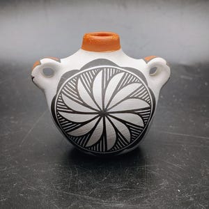 May include: A small ceramic vessel with a white base, featuring a black circular geometric pattern of radiating petals on the front. The piece has a terracotta-colored rim and two side handles, with additional terracotta accents on the handles.