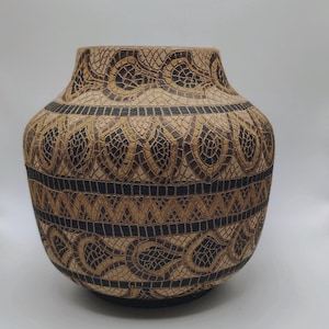 May include: A decorative, handcrafted vase with a wide base and a narrower neck. The vase features intricate, mosaic-like patterns in shades of brown and black. The design includes repeating geometric and floral motifs, creating a textured appearance.
