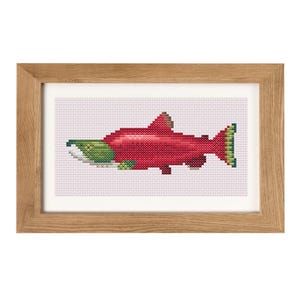 May include: Cross-stitch artwork of a red salmon with green details, presented in a light wood frame. The fish is rendered in a pixelated style, set against a white backdrop. The frame is a natural wood colour.