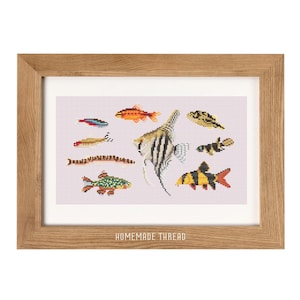 May include: A framed cross-stitch artwork featuring various colorful fish designs on a light pink background. The artwork is in a light brown wooden frame. The text "HOMEMADE THREAD" is at the bottom of the artwork.