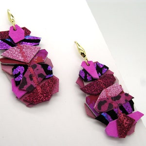 Puede incluir: Un par de pendientes colgantes de color rosa, morado y negro con un gancho dorado. Los pendientes están hechos de cuero y purpurina en capas.
