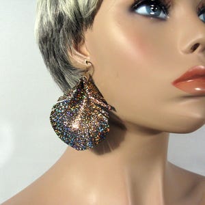 May include: A pair of large, iridescent feather earrings. The earrings are made of a metallic material with a textured surface. The earrings are a mix of gold, brown, and blue colours.
