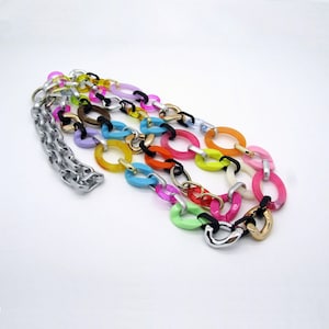 Puede incluir: Un collar multicadena colorido con eslabones grandes, gruesos y ovalados en varios colores brillantes, incluyendo rosa, naranja, amarillo, verde, azul y morado. El collar está hecho de plástico y tiene una cadena plateada unida a él.