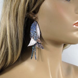 Può includere: Un paio di orecchini lunghi e pendenti con un design a piuma blu, rosa e bianco. Le piume sono decorate con glitter iridescente.