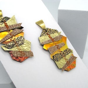 May include: A pair of gold and orange geometric earrings with a glittery finish. The earrings are made of layered pieces of leather and have a unique, modern design.