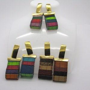 Puede incluir: Seis pares de pendientes de cuero a rayas y coloridos con ganchos dorados. Cada pendiente presenta una combinación de colores diferente de tiras de cuero.