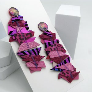 Puede incluir: Un par de pendientes geométricos vibrantes en tonos rosa, magenta y morado. Los pendientes presentan formas en capas con detalles de purpurina y estampado animal, y una parte superior circular con motas doradas. El diseño es audaz y llamativo.