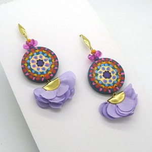 May include: A pair of dangle earrings featuring a colourful mandala design on a teal background. The mandala is surrounded by a gold trim and a layer of purple fabric flowers.