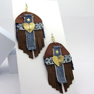 Puede incluir: Un par de pendientes de cuero marrón con flecos, mezclilla y detalles dorados. Cada pendiente presenta un diseño en forma de corazón con una estrella dorada y un corazón dorado.