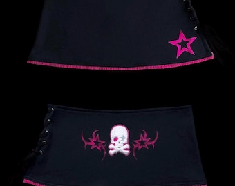 Puede incluir: Una falda negra con un diseño de estrella rosa y cordones laterales. El dobladillo tiene costuras rosas. Una segunda vista muestra un diseño de calavera blanca flanqueado por estrellas rosas, también con cordones laterales y dobladillo rosa.