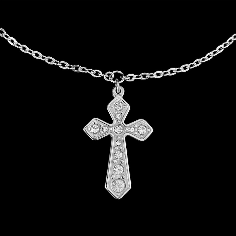 May include: A silver-colored cross pendant necklace with clear, sparkling gemstones. The cross is suspended from a delicate chain against a black background. The cross design features a classic Christian symbol.