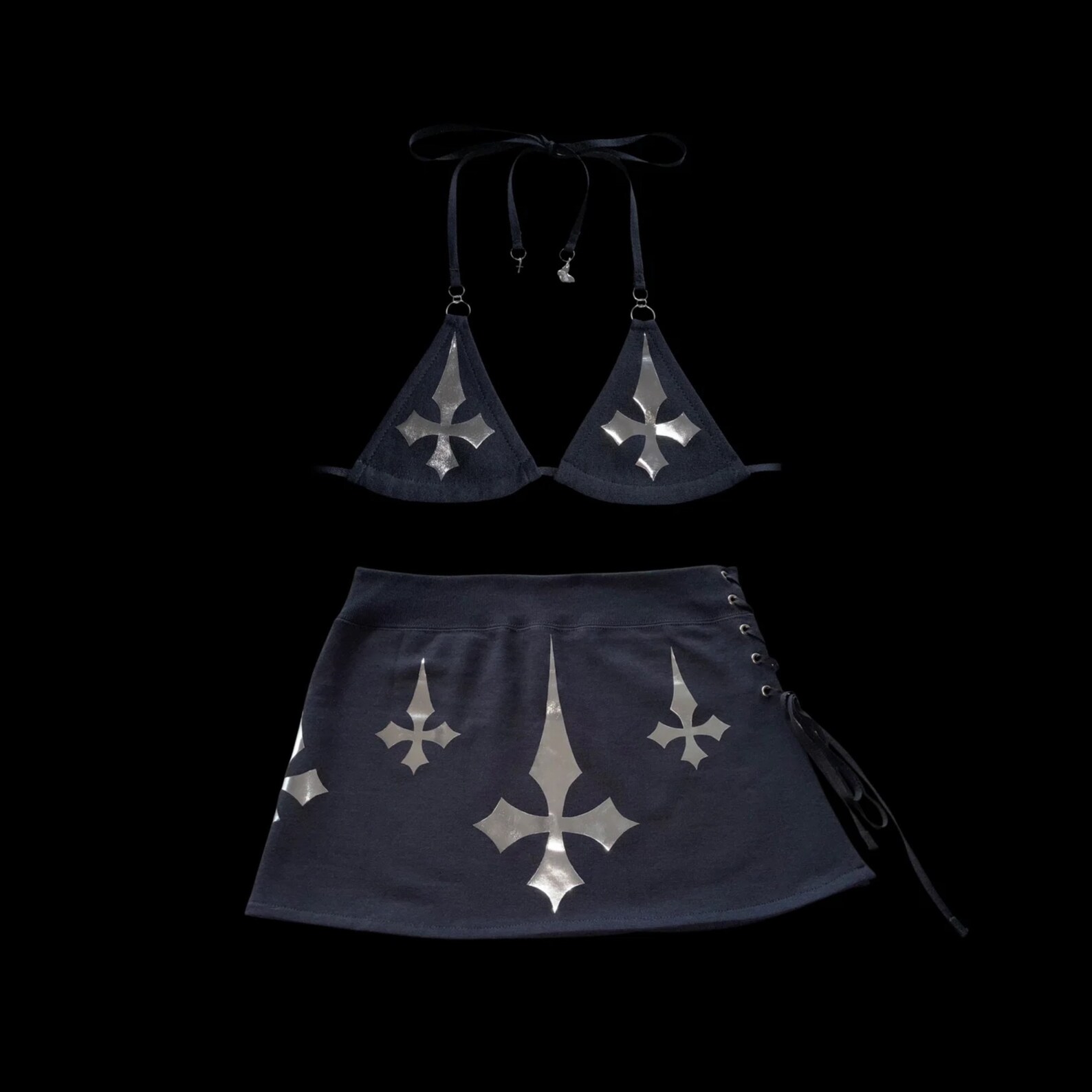 Flat lay of the Unholy two-piece set with black triangle bikini top and matching mini skirt, both featuring silver inverted cross designs.