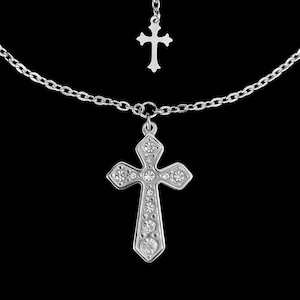May include: A silver-toned necklace featuring a large cross pendant adorned with clear crystals. A smaller cross hangs above the main pendant, connected by a delicate chain. The necklace is set against a black background.