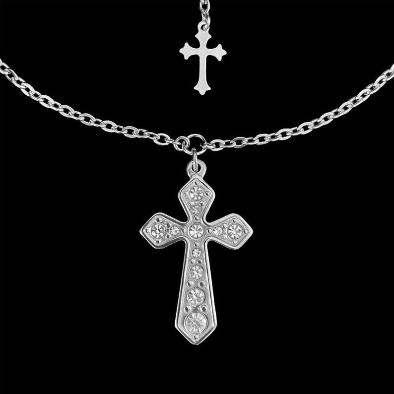 May include: A silver-toned necklace featuring a large cross pendant adorned with clear crystals. A smaller cross hangs above the main pendant, connected by a delicate chain. The necklace is set against a black background.