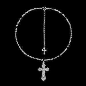 May include: A silver-colored necklace with two cross pendants. The larger cross is embellished with small, clear stones. The necklace has a delicate chain design and a dangling cross pendant. The background is solid black.