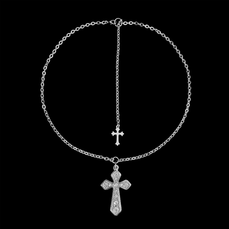 May include: A silver-colored necklace with two cross pendants. The larger cross is embellished with small, clear stones. The necklace has a delicate chain design and a dangling cross pendant. The background is solid black.