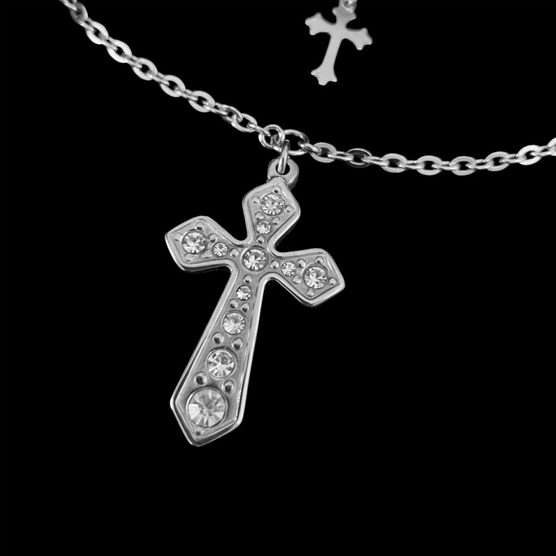May include: A silver-colored necklace with a cross pendant. The cross is adorned with clear, sparkling gemstones. The chain is a delicate link style, and a smaller cross charm is also visible.