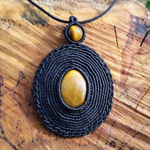 May include: A handcrafted macrame necklace featuring a black woven pendant with an oval, golden-brown gemstone at its center. The pendant is suspended from a black cord, showcasing a smaller gemstone at the top. The necklace rests on a wooden surface.