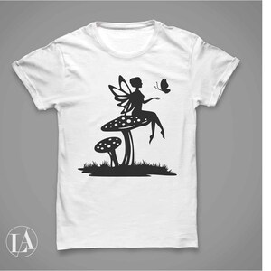 Puede incluir: Camiseta blanca con una silueta negra de un hada sentada en un hongo con una mariposa volando cerca.