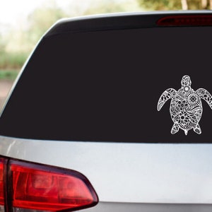 Puede incluir: Una calcomanía de vinilo blanco con un diseño de tortuga detallada y adornada en la ventana trasera de un coche negro.