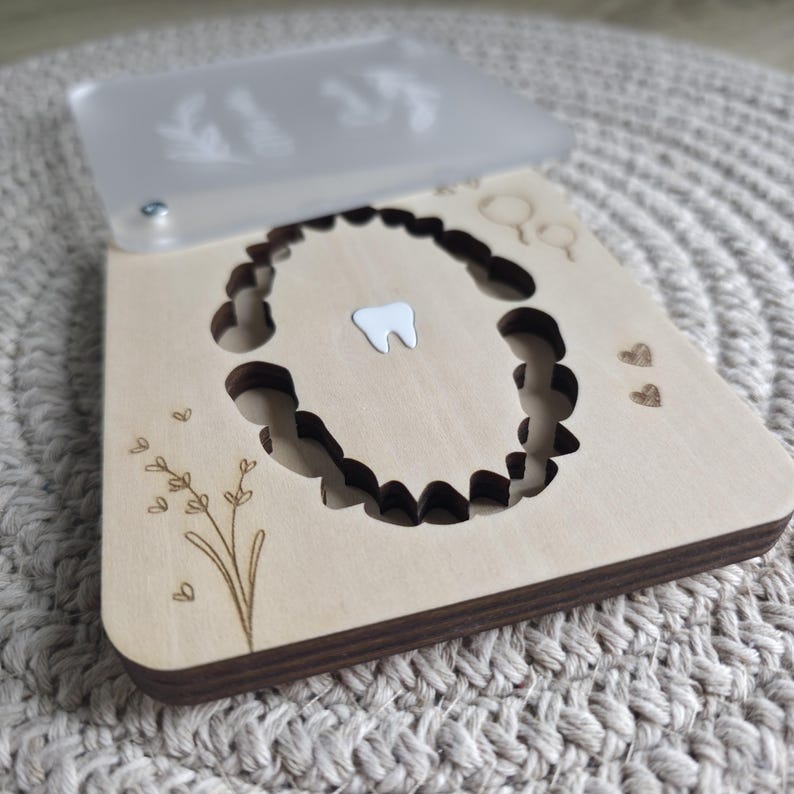 Puede incluir: Una caja de madera para el hada de los dientes con tapa transparente. La caja presenta un recorte de una boca con dientes y un solo diente blanco. La tapa tiene un dise&ntilde;o floral. La caja est&aacute; decorada con corazones grabados y una ramita floral.