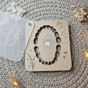 Puede incluir: Un cuadro de dientes de madera con un recorte de boca y un solo diente blanco. El cuadro tiene detalles grabados de globos, corazones y elementos florales. Se adjunta una cubierta transparente y esmerilada con el texto "Mes petites".