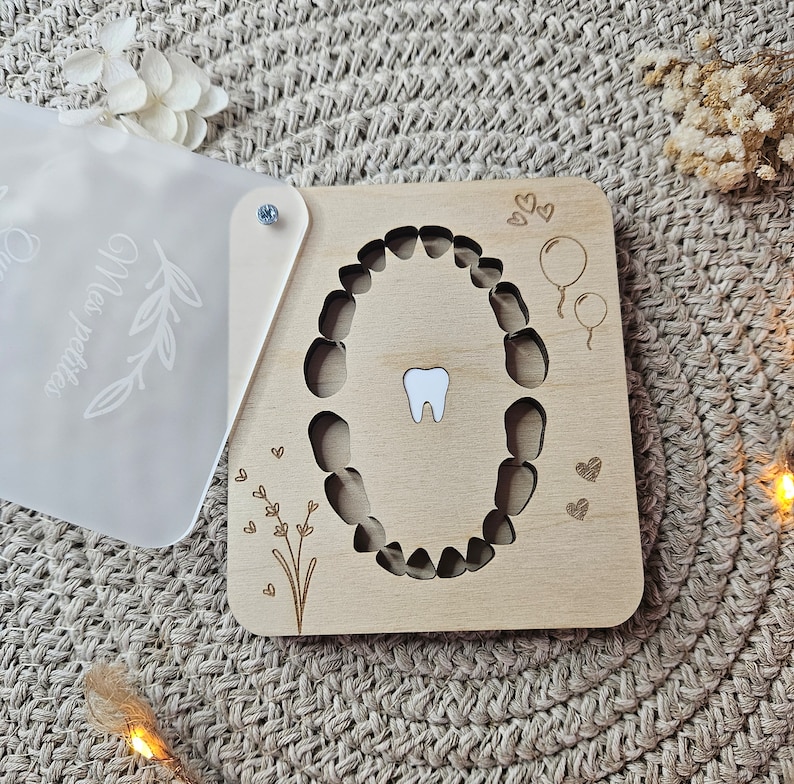 Puede incluir: Un cuadro de dientes de madera con un recorte de boca y un solo diente blanco. El cuadro tiene detalles grabados de globos, corazones y elementos florales. Se adjunta una cubierta transparente y esmerilada con el texto "Mes petites".
