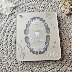 Puede incluir: Una caja de recuerdos rectangular de madera con una cubierta de acr&iacute;lico transparente. La caja presenta un dise&ntilde;o en forma de diente con las palabras "Mes petites Quenottes" y el nombre "Chloe". Los elementos decorativos incluyen corazones, globos y acentos florales.