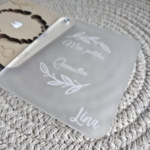 Puede incluir: Un letrero rectangular esmerilado con las palabras "Mes petites Quenottes Lina" en escritura blanca. El letrero tiene un peque&ntilde;o remache plateado y est&aacute; colocado sobre una superficie gris texturizada. Un objeto de madera es parcialmente visible en el fondo.