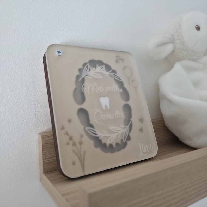 Puede incluir: Una caja de madera beige para el hada de los dientes con una parte frontal de acr&iacute;lico transparente. La caja presenta el texto "Mes petites Quenottes" y un gr&aacute;fico de un diente. La caja est&aacute; personalizada con el nombre "Lina". Un cordero de peluche blanco est&aacute; en el fondo.