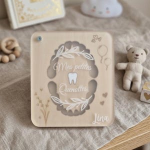 Puede incluir: Una caja de madera de color claro para el hada de los dientes con el texto "Mes petites Quenottes" y el nombre "Lina". La caja presenta un recorte en forma de diente con un gr&aacute;fico de diente, dise&ntilde;os florales y de coraz&oacute;n. Un peque&ntilde;o oso de peluche y otros elementos decorativos est&aacute;n en el fondo.