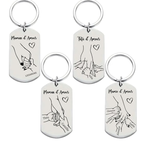 May include: Four silver keychains with a dog tag design. Each keychain features a black and white illustration of two hands holding each other. The text "Maman d'Amour", "Tata d'Amour", "Maman d'Amour", and "Mamie d'Amour" is printed on each keychain. The names of children are printed below the illustration on each keychain.
