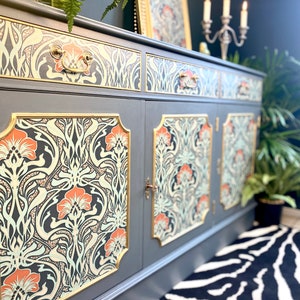 May include: A dark grey sideboard with two doors and three drawers. The doors and drawers are decorated with a floral pattern in shades of orange, green, and black. The sideboard has gold trim and brass handles.