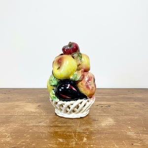 Può includere: Una scultura di cestino di frutta in ceramica con una varietà di frutta colorata. La scultura comprende mele, prugne e altri frutti nei toni del rosso, giallo e viola. Il cestino è bianco con un design intrecciato.