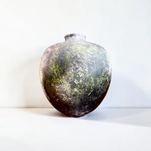 Puede incluir: Un jarrón de cerámica redondeado en forma de corazón con una superficie texturizada y moteada. El acabado presenta una mezcla de tonos gris oscuro, negro y verde musgo, creando una apariencia desgastada con una pequeña abertura circular.