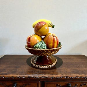 Può includere: Una fruttiera in ceramica con una banana e pesche. Le pesche sono rosse e gialle, e la banana è gialla. La ciotola è marrone con una trama intrecciata e la base è marrone più scuro. È inclusa anche una foglia verde.