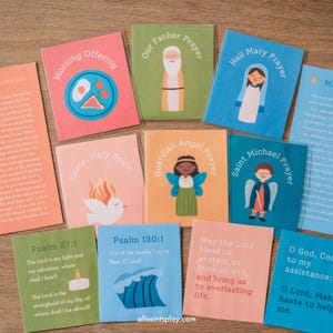 Può includere: Una collezione di colorate preghiere illustrate con testo. Le carte presentano varie preghiere, tra cui "Padre Nostro" e "Ave Maria". Le carte sono disposte su una superficie di legno.