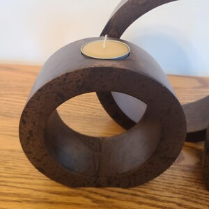 Op de afbeelding: Een donkerbruine houten kandelaar met een cirkelvormig ontwerp. Een kleine, ronde kaars zit in een metalen houder in de bovenste cirkel. Het hout heeft een textuur en het ontwerp is modern en minimalistisch.